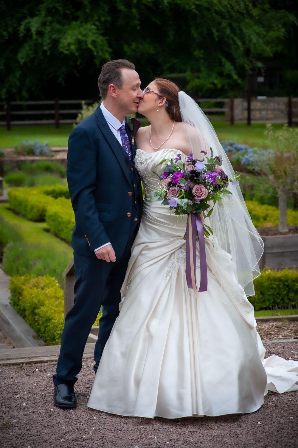 Wedding couple shares intimate moment in landscaped garden with purple floral bouquet.