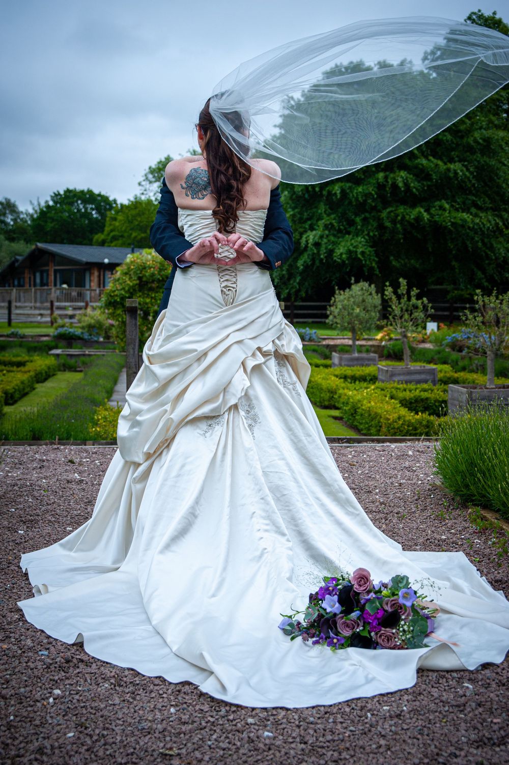Back view of elegant white wedding dress with flowing train and purple bouquet in formal garden.