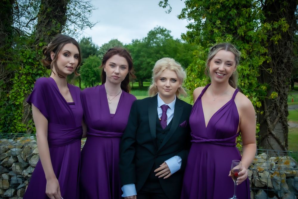 Wedding party poses outdoors wearing coordinated purple dresses and dark suit.