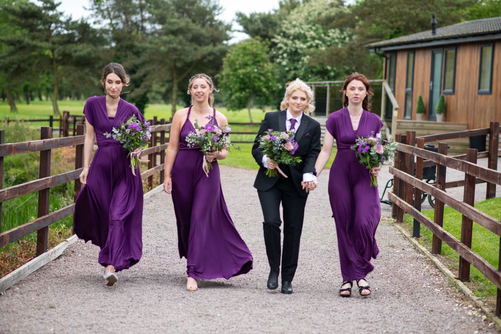 Wedding party walks along countryside path wearing vibrant purple dresses.