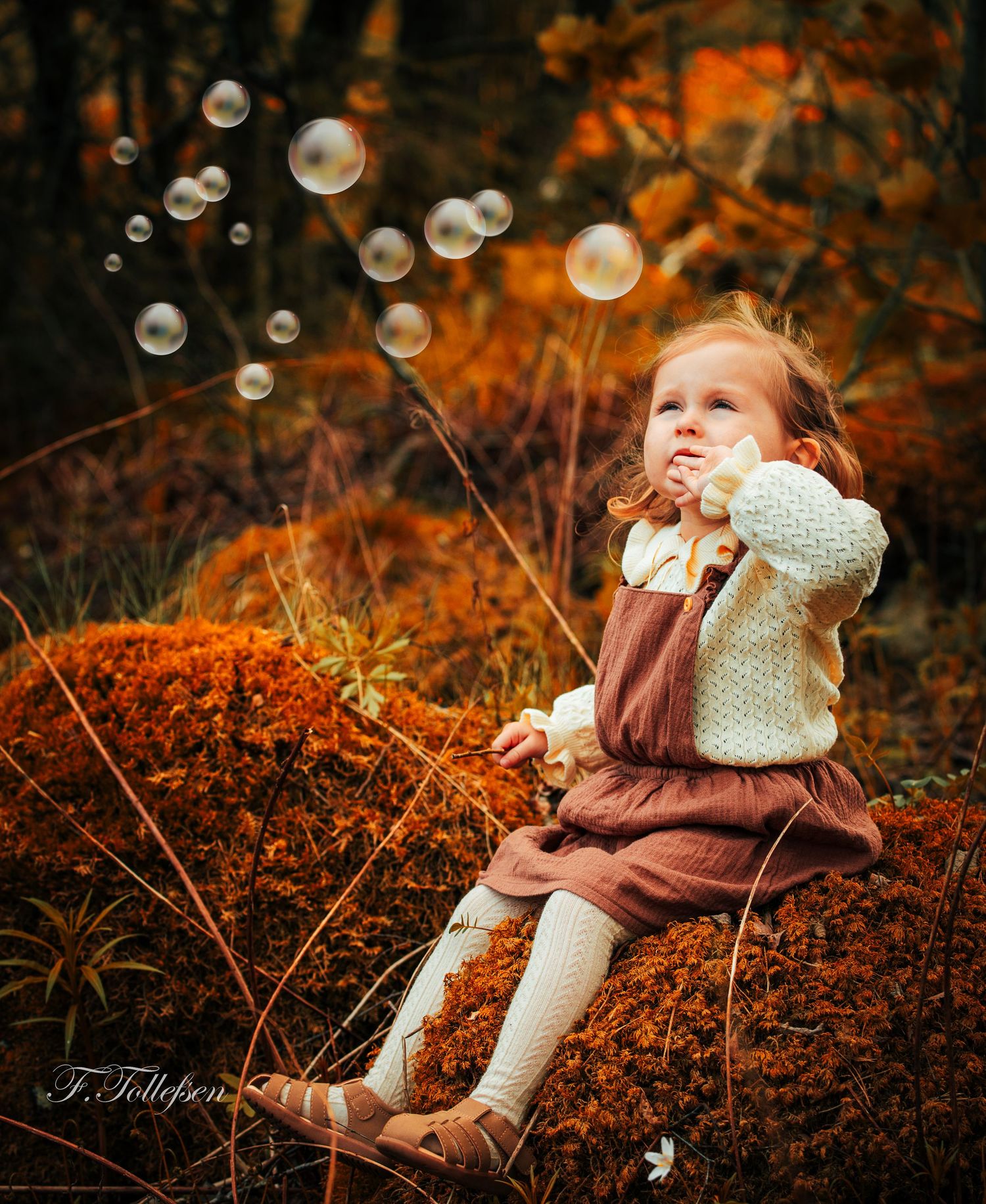 Young girl in autumn forest blowing soap bubbles while sitting on mossy rocks.