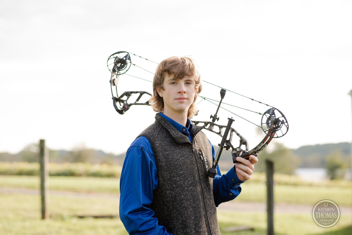 Person in blue and gray vest holds compound bow while standing in rural setting.