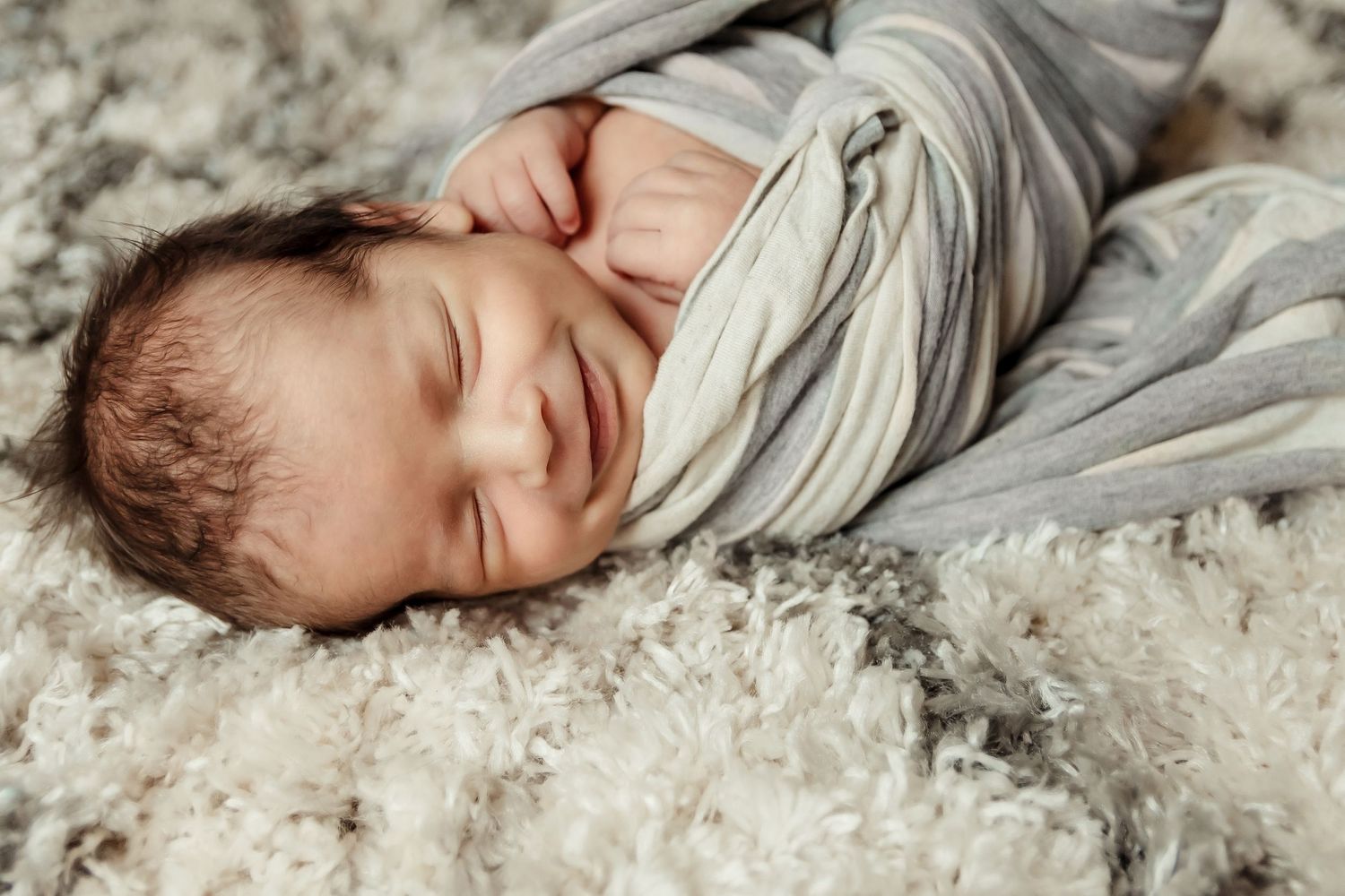 Sleeping newborn baby wrapped in grey swaddle lying on soft white furry blanket.