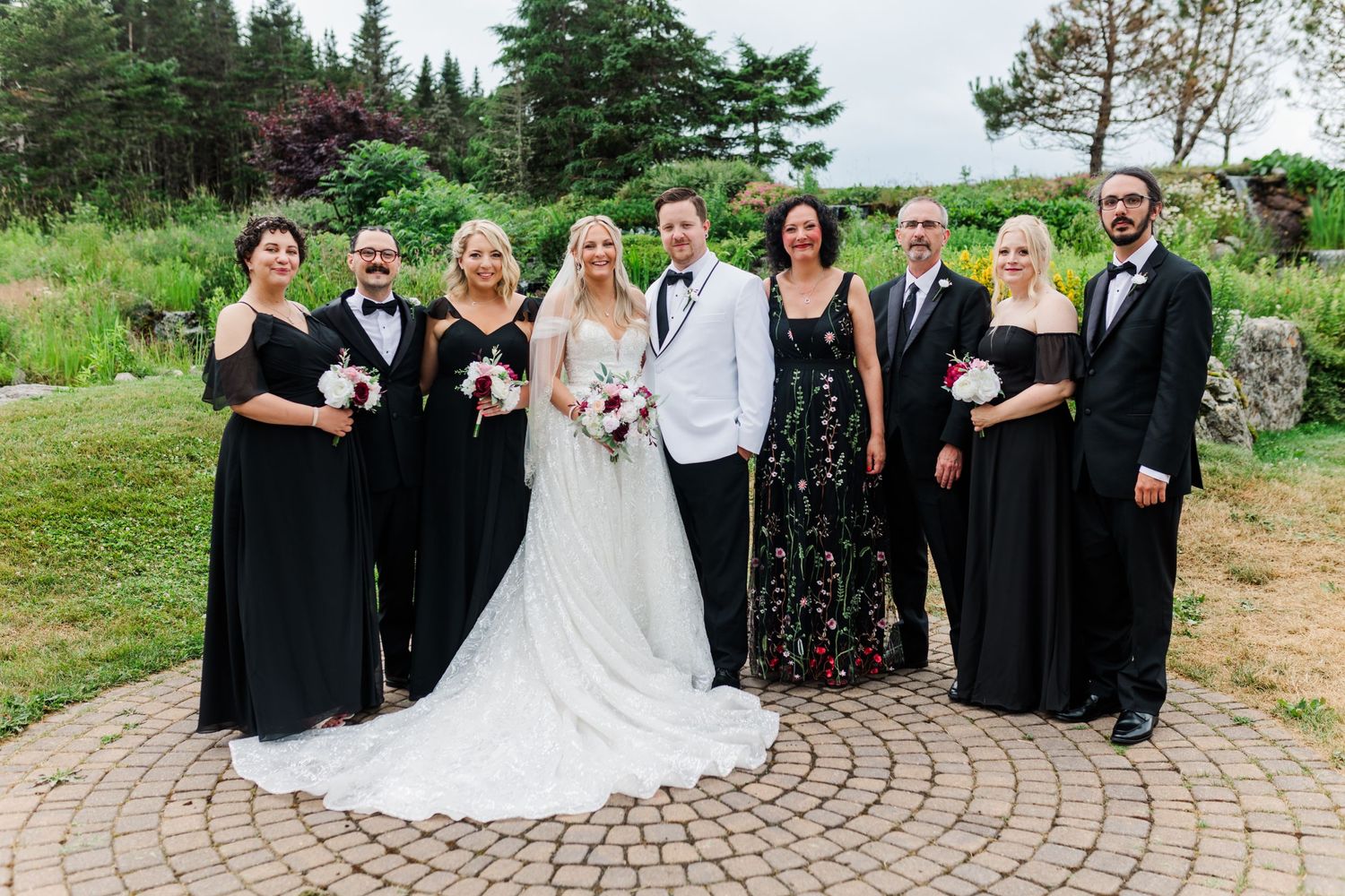 Wedding party poses in formal attire on circular stone patio surrounded by garden landscape.