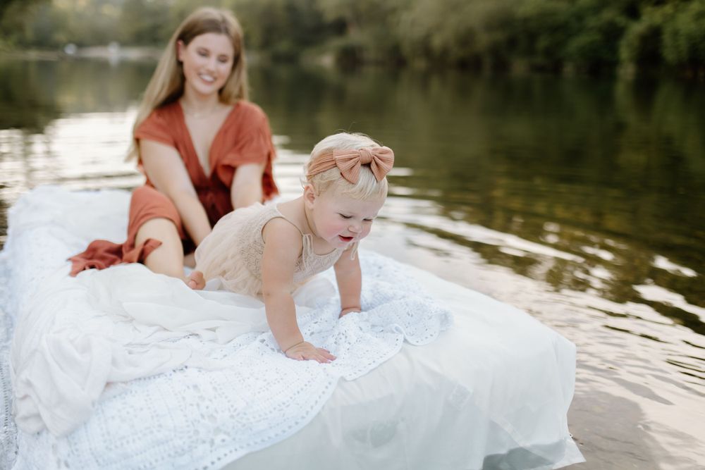 Mom and baby on an air mattress covered in white lace fabric while floating in river. baby crawling to edge to see water