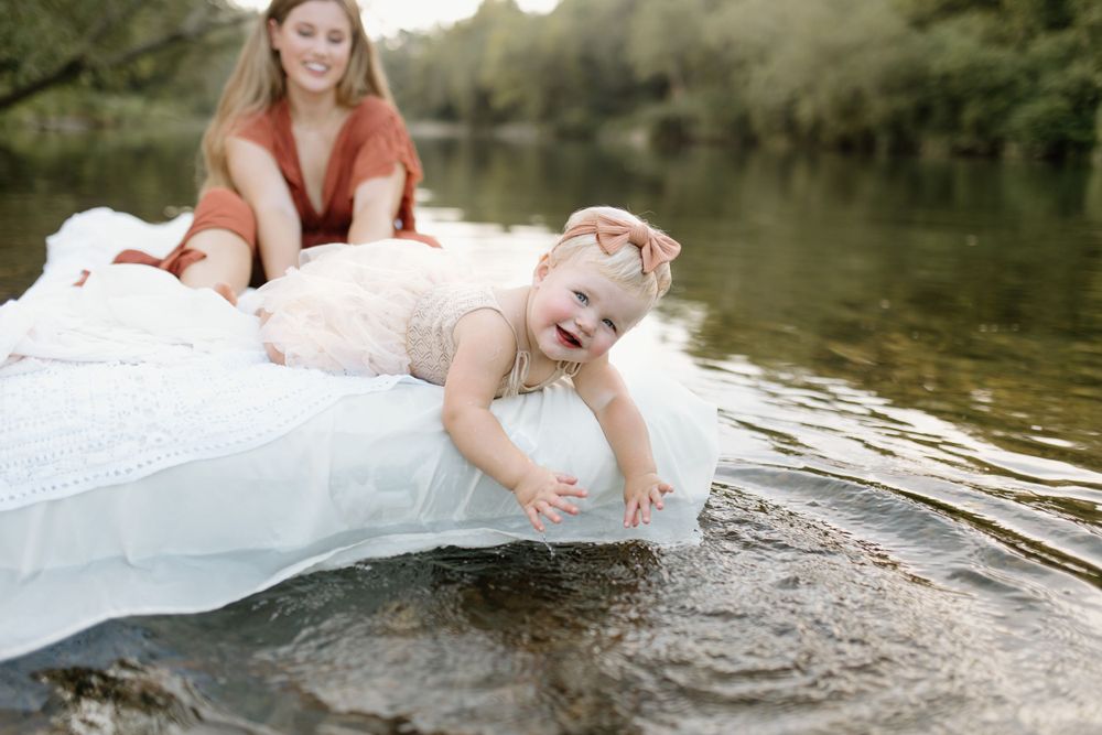 Mom and baby on an air mattress covered in white lace fabric while floating in river. baby leaned over splashing water