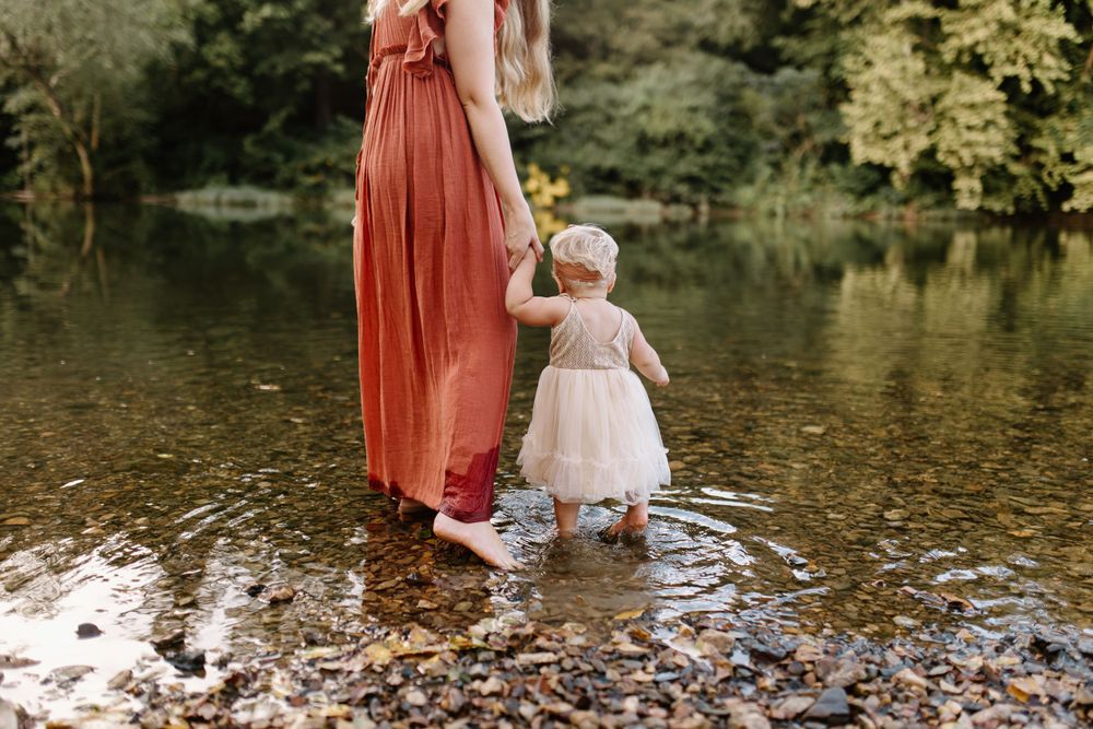Mom and baby holding hands walking into a shallow pebble bottomed river