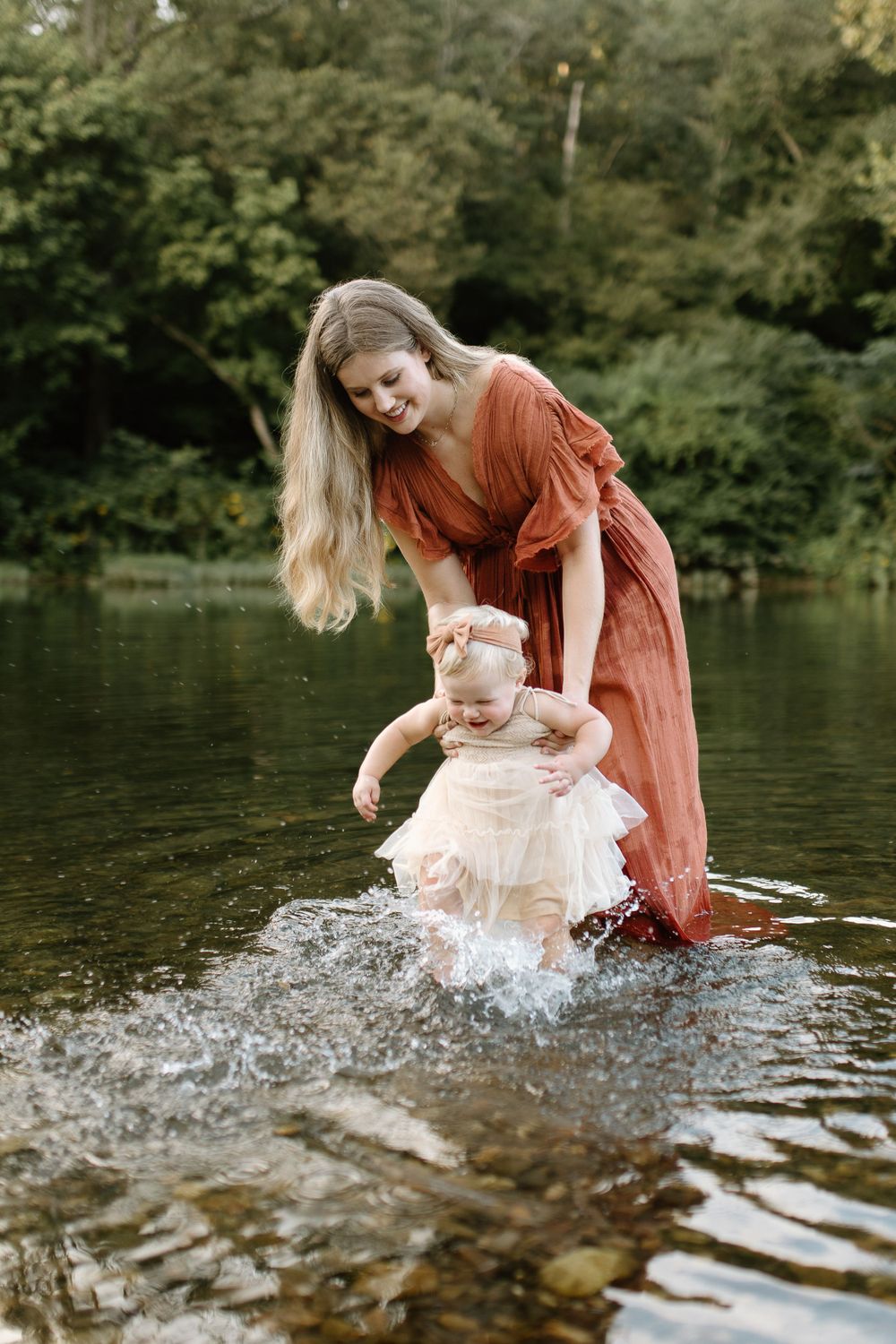 Mom holding baby girl slightly above the water as baby kicks feet splashing in the river