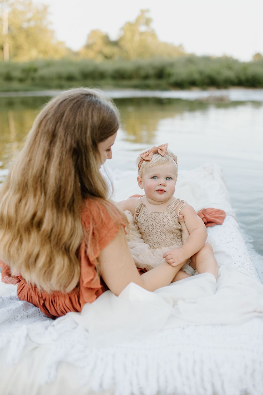 Mom and baby girl snuggling on an air mattress covered in white lace fabric while floating in shallow river
