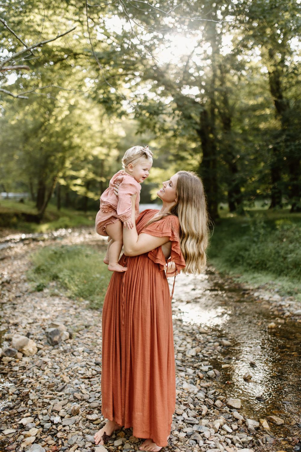 Momma and baby girl play in a small pebble bottomed creek as the dreamy sunlight filters through the surrounding trees