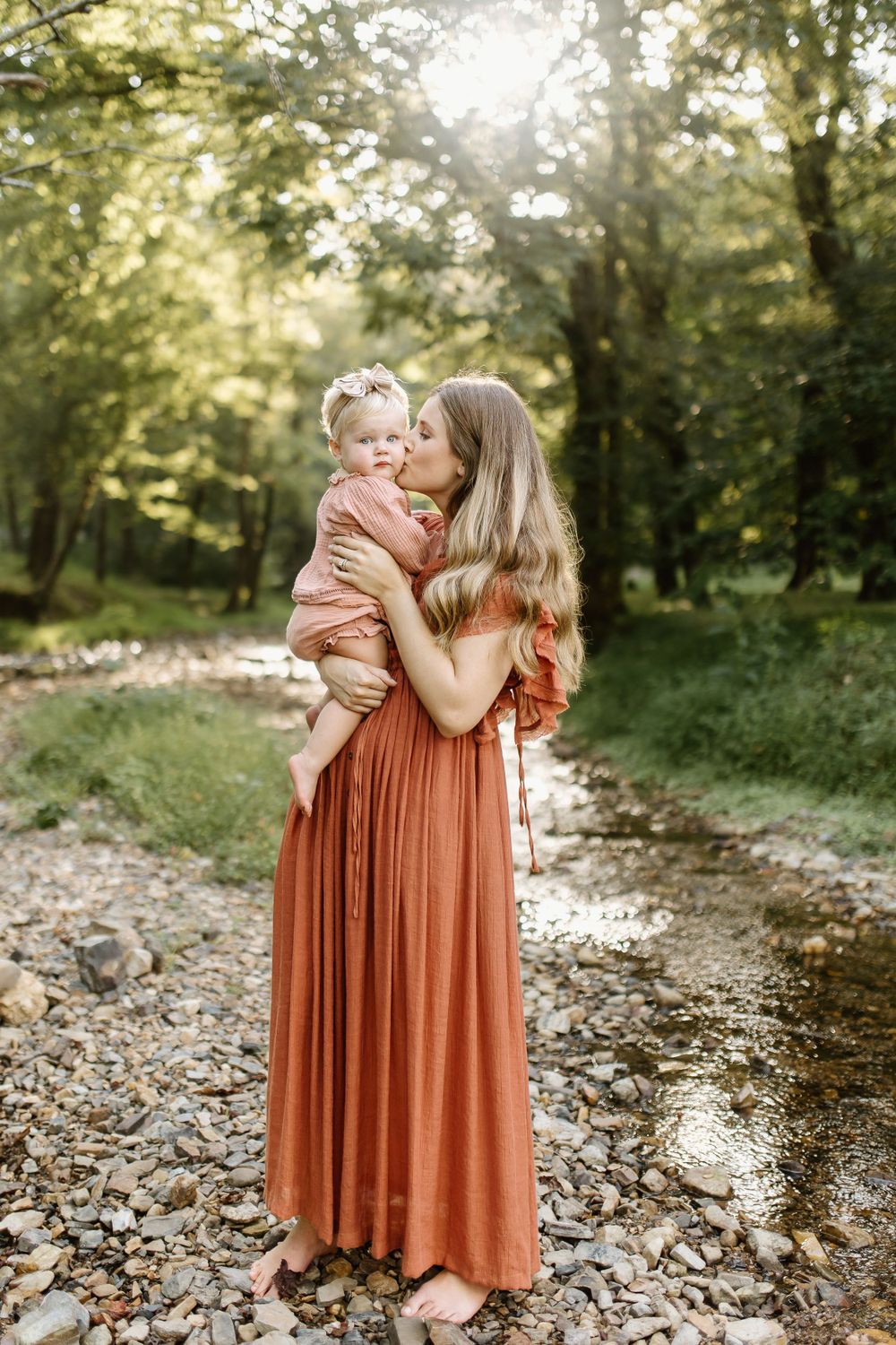 Momma and baby girl play in a small pebble bottomed creek as the dreamy sunlight filters through the surrounding trees
