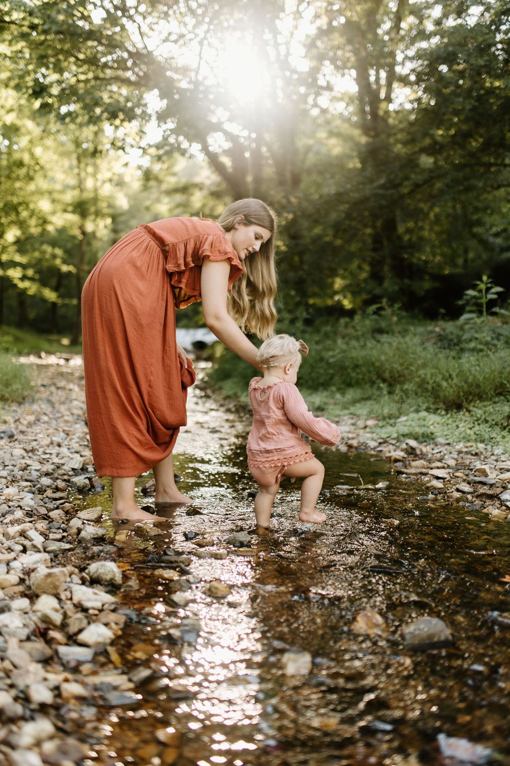 Momma and baby girl play in a small pebble bottomed creek as the dreamy sunlight filters through the surrounding trees