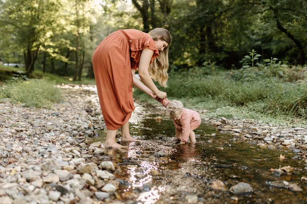 Momma and baby girl play in a small pebble bottomed creek as the dreamy sunlight filters through the surrounding trees