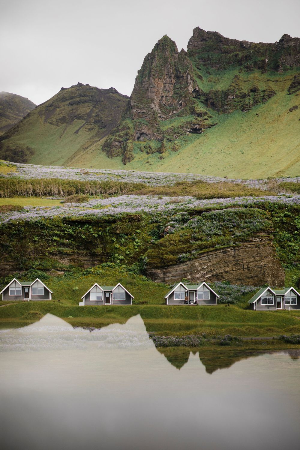 Small white cottages reflect in calm water below dramatic green mountain peaks in Iceland's rugged landscape.