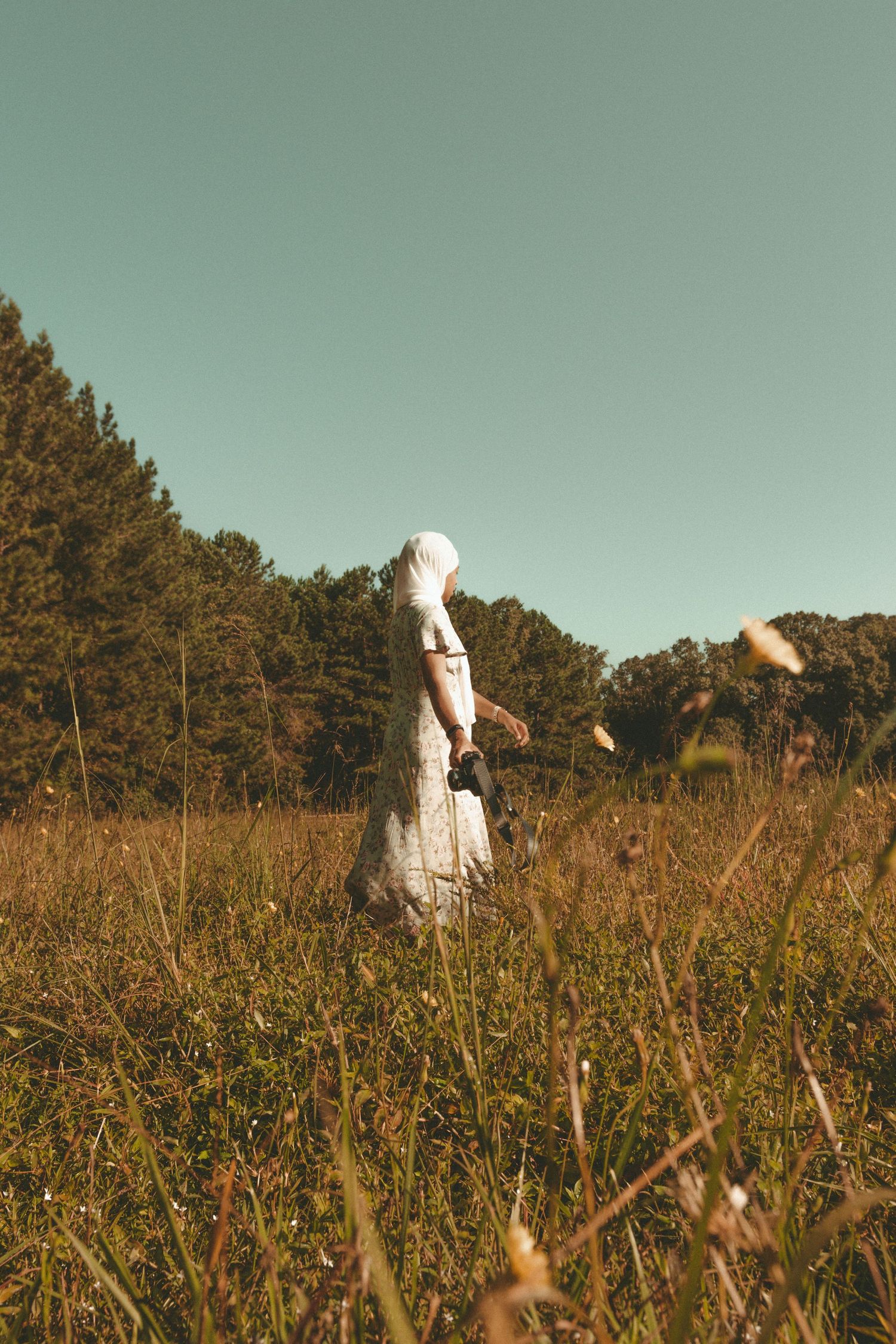 Person in white clothing walking through a tall grass field against a teal sky.