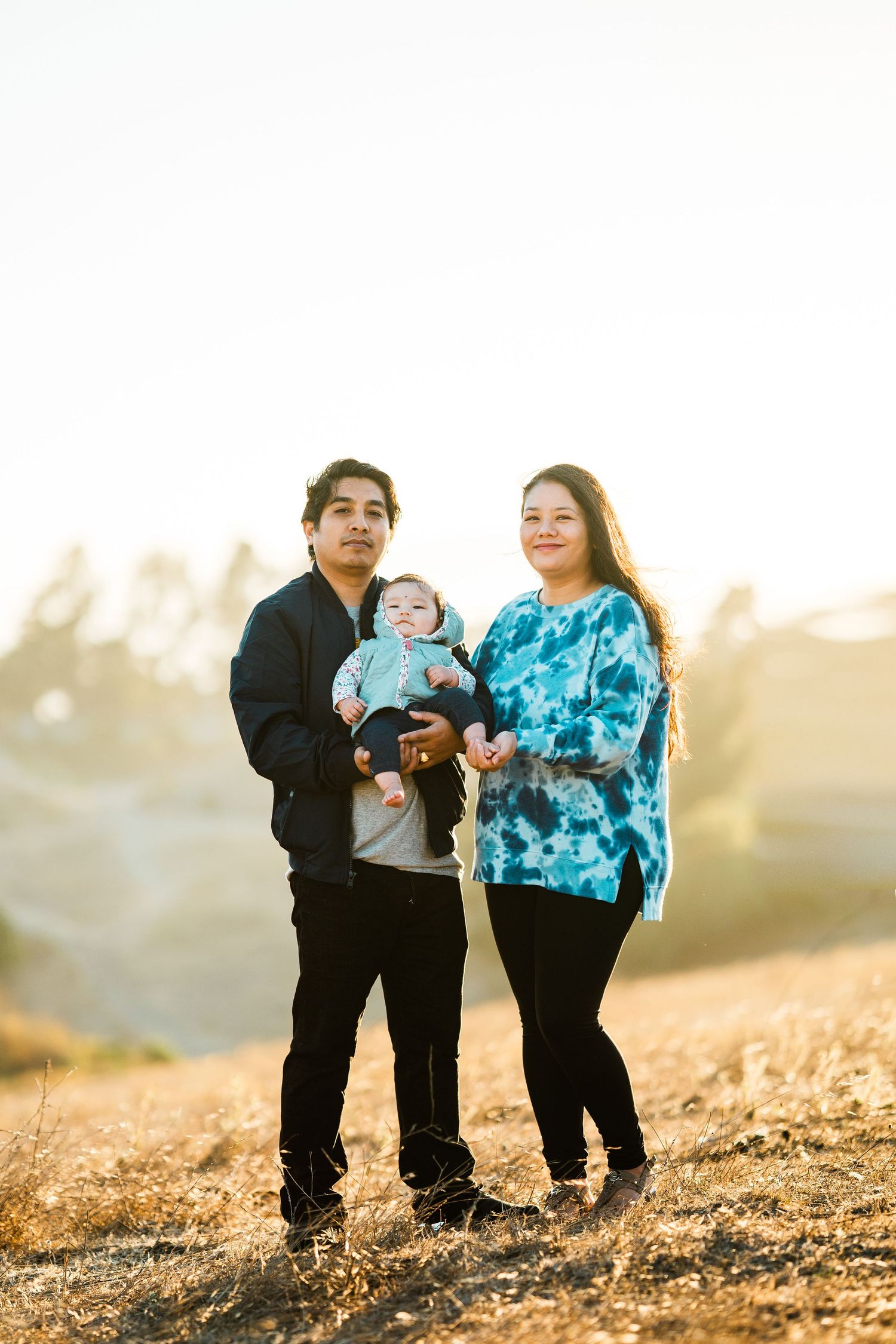 Family of three stands together on a golden hillside during sunset wearing casual clothing.