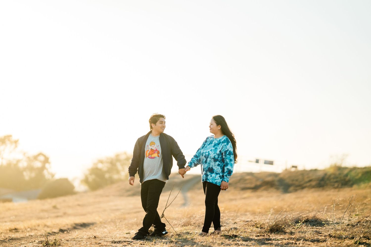 Couple holding hands while walking together on a misty hilltop at sunset in casual attire.
