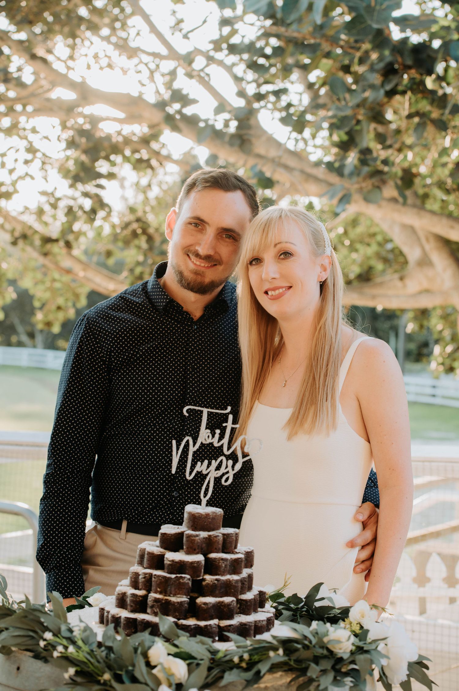 A couple stands behind their tiered chocolate dessert display at an outdoor event.