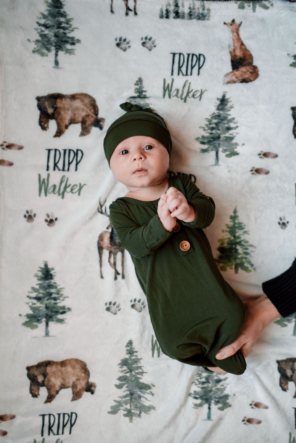 Baby wearing green outfit on woodland-themed blanket with bear and deer prints.