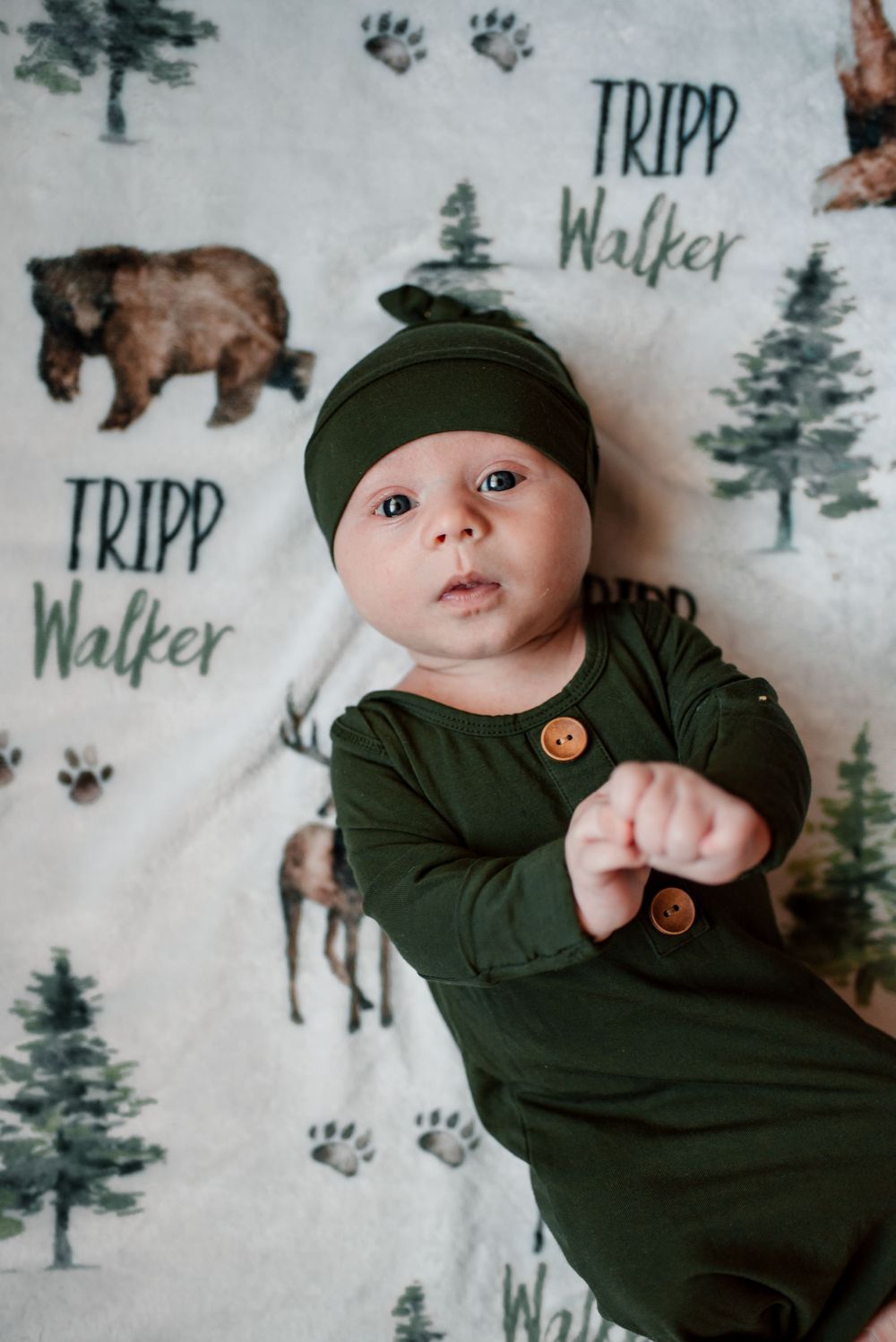 Baby in dark green outfit and beanie on woodland-patterned blanket.