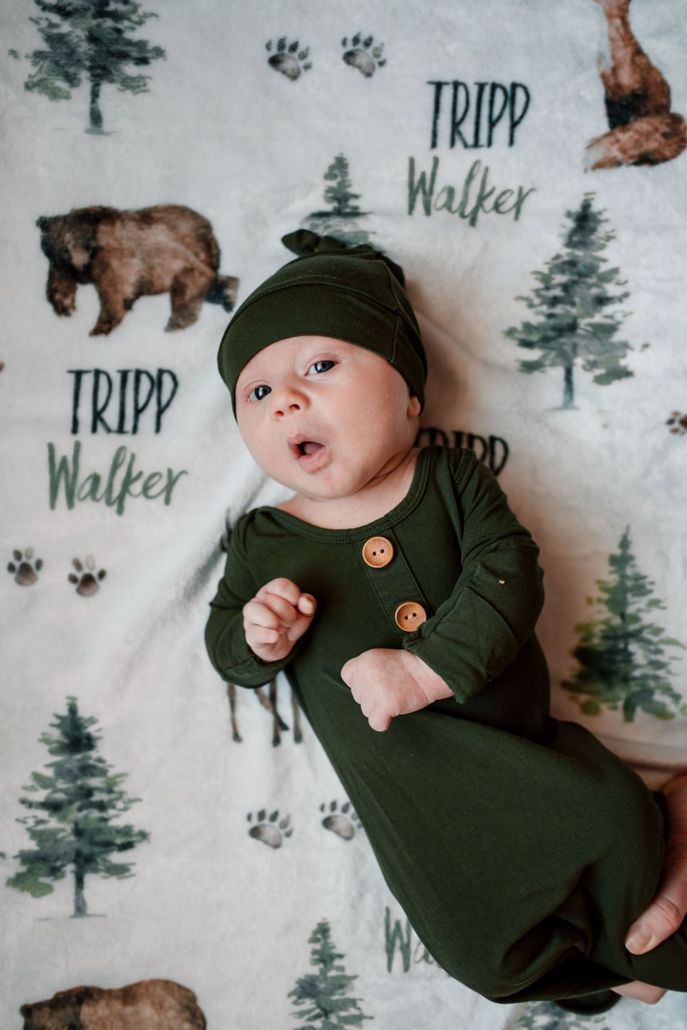 Baby in green sleeper and beanie lying on woodland-themed blanket with animal prints.
