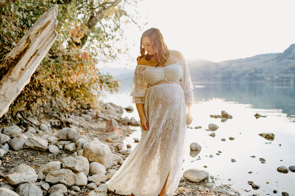 Sunlit maternity photos by a serene lake with mountains and rocks creating a peaceful outdoor setting.