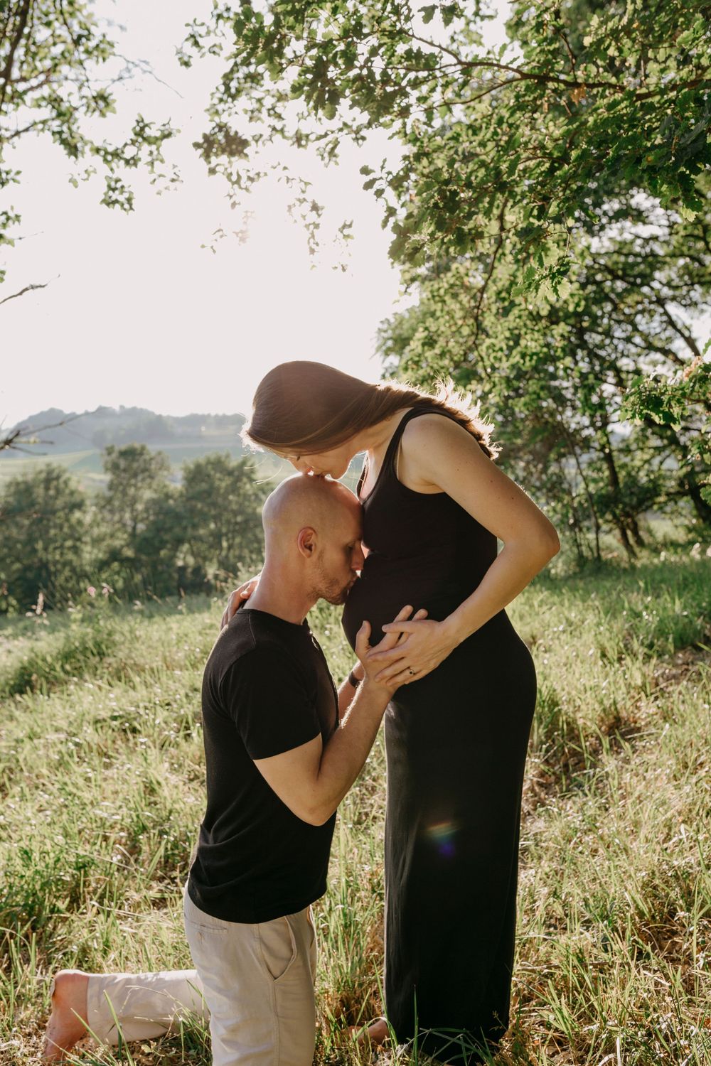 A couple embraces tenderly in a sunlit meadow surrounded by lush greenery.