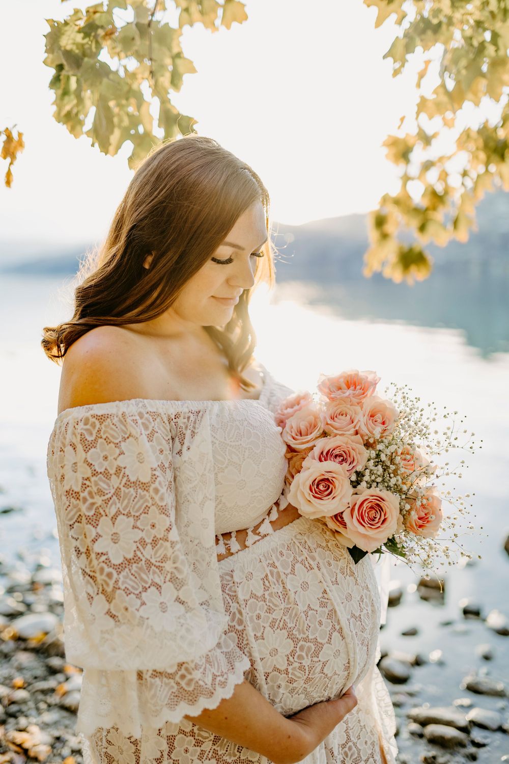 Dreamy maternity portraits featuring a white lace dress and pink roses against a lakeside backdrop.