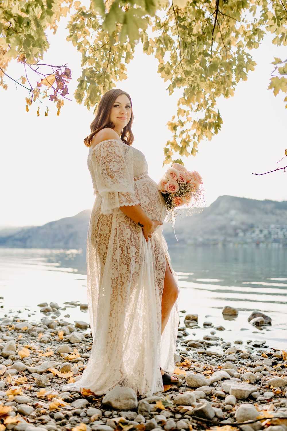 Elegant maternity photography on a rocky shoreline with mountains and autumn foliage framing the scene.