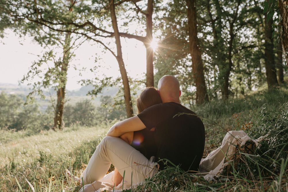 A romantic couple shares an intimate moment under sunlit trees in a scenic outdoor setting.