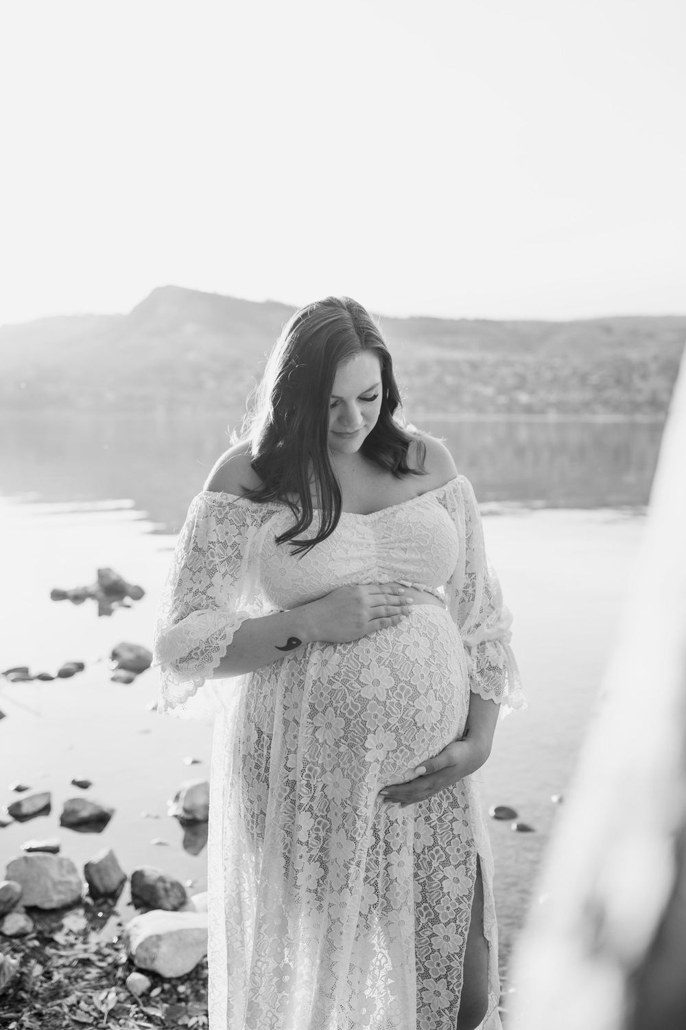 A romantic maternity photoshoot on a rocky beach shore with a white lace dress and mountains in the background.