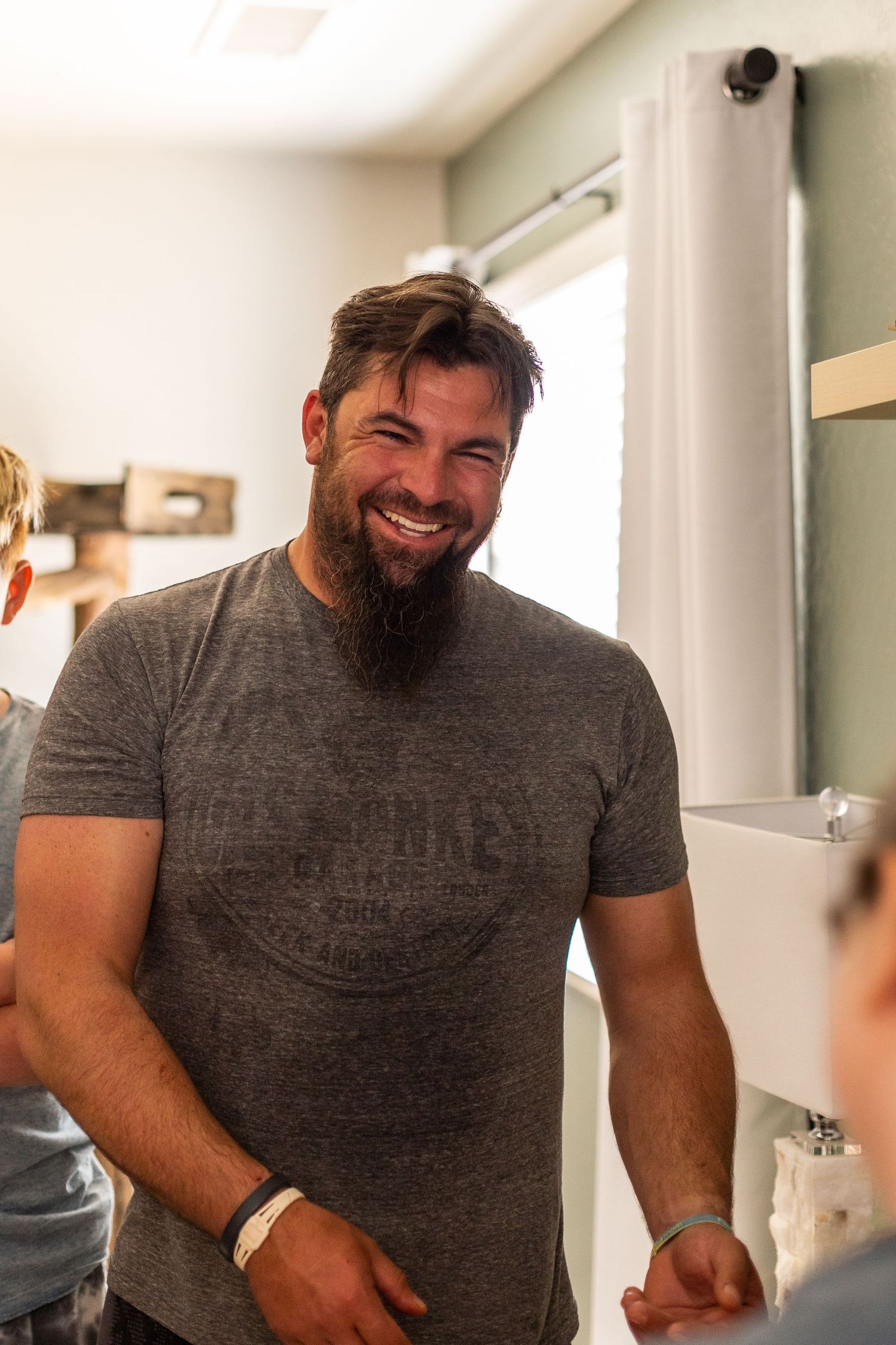 A bearded man in a gray t-shirt smiles warmly while standing in a bright indoor setting.