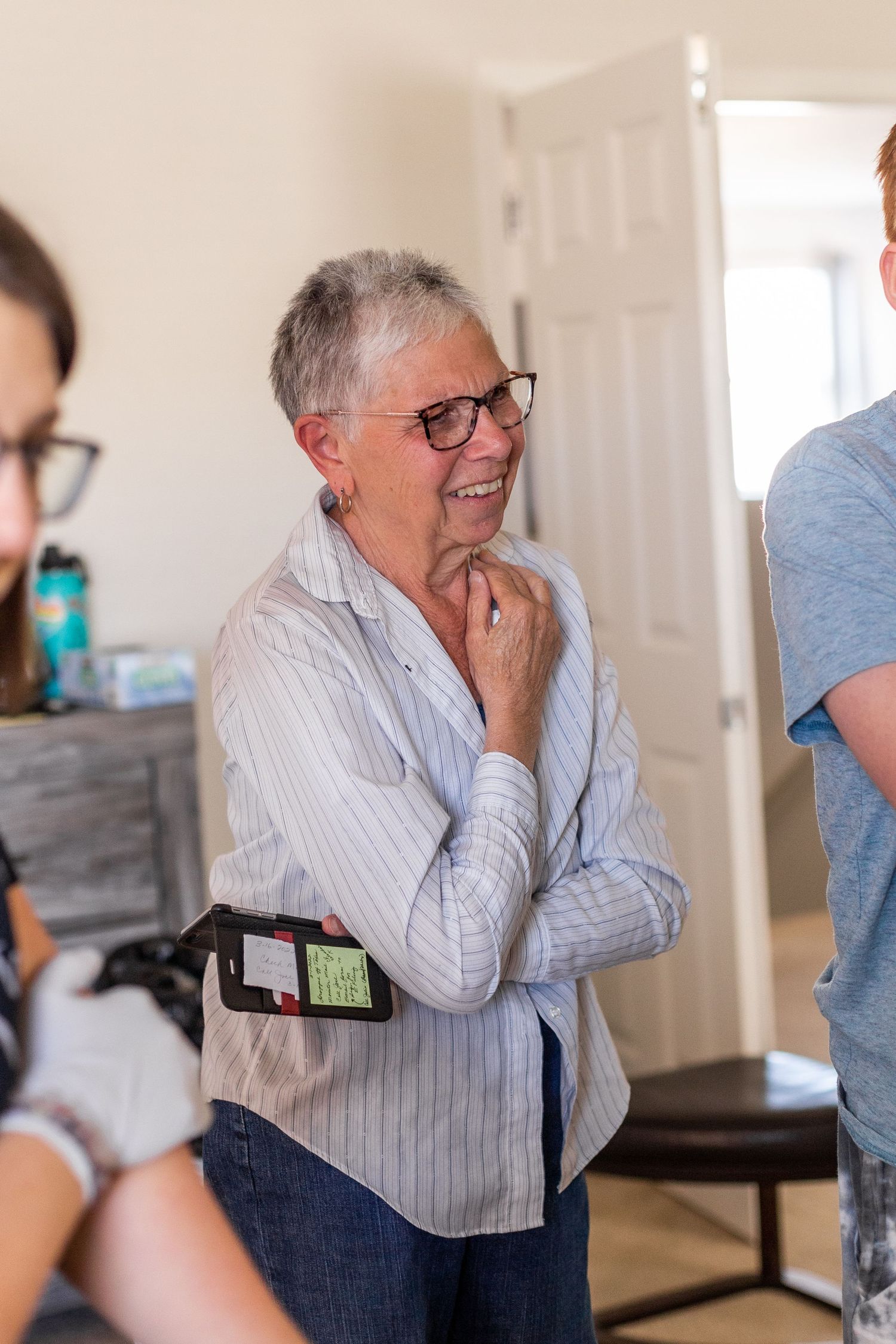 An elderly person in glasses and striped shirt stands with a thoughtful expression indoors.