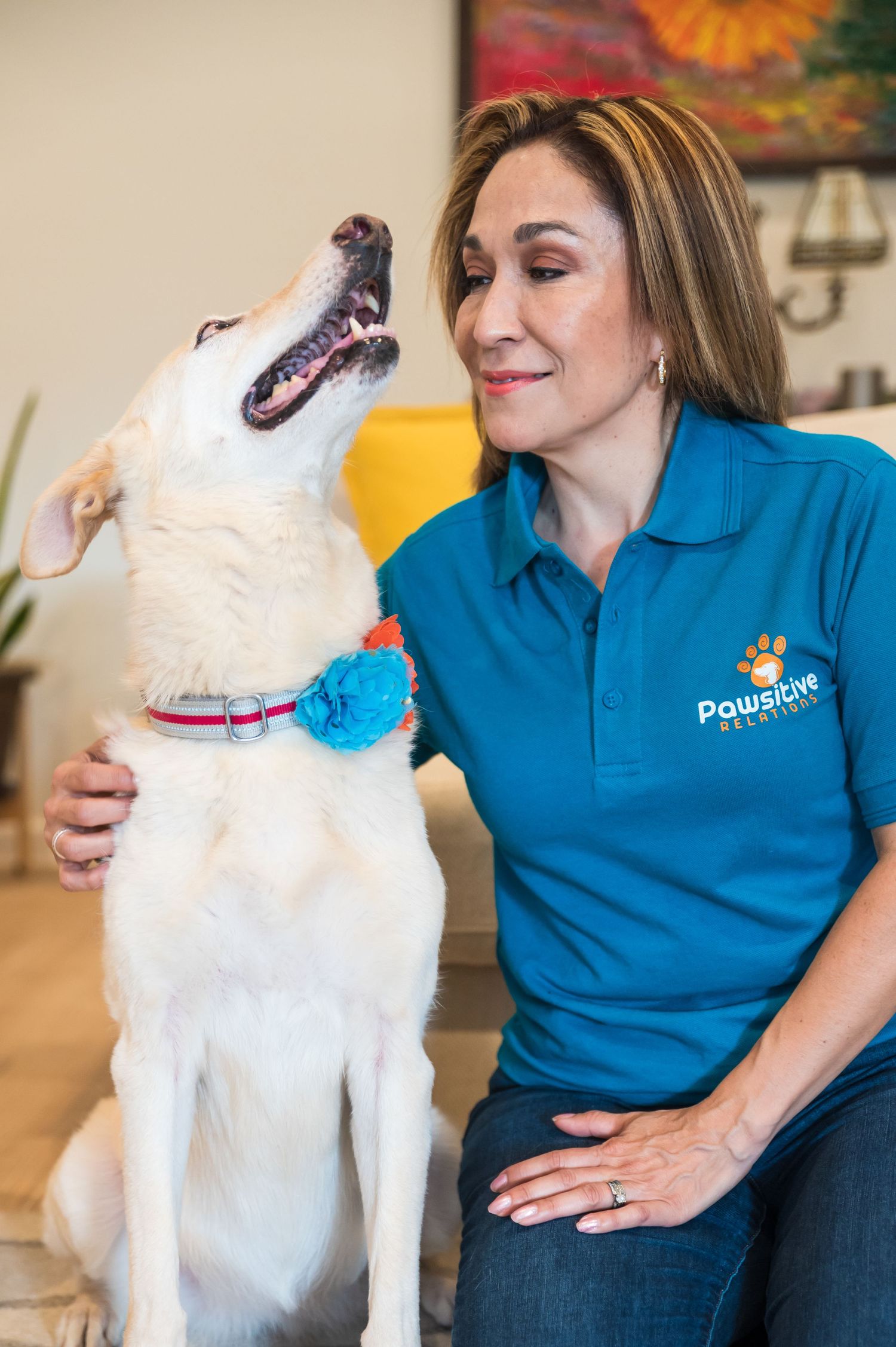 A white dog wearing a blue flower collar accessory interacts with a professional pet care provider.