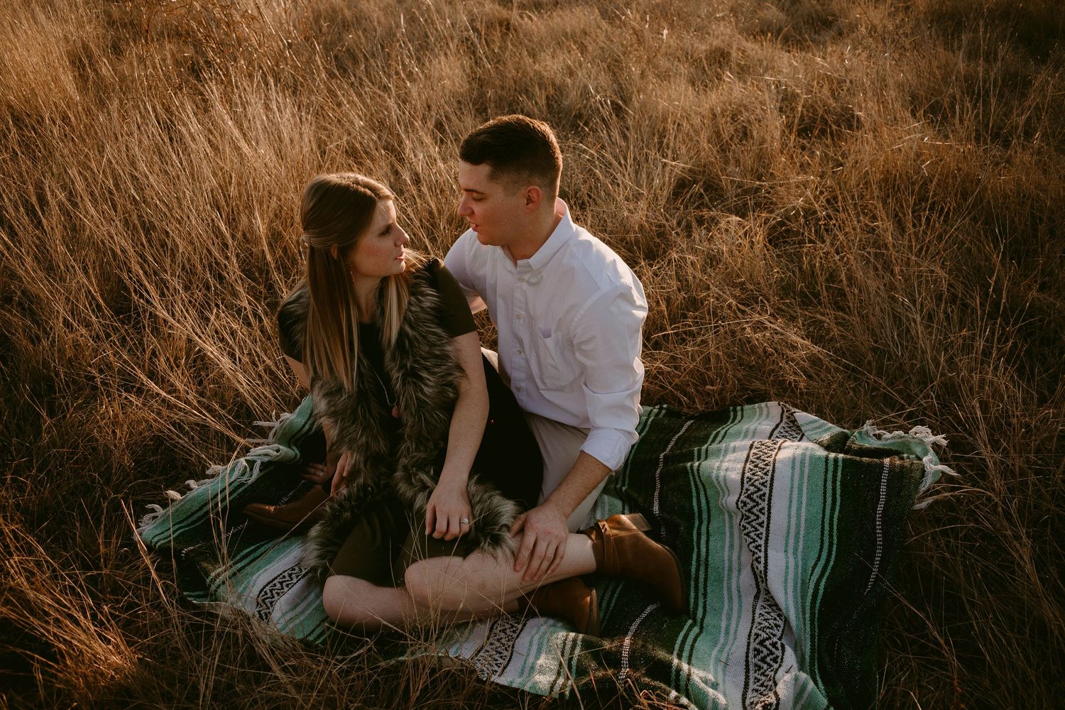 A romantic couple sits on a striped blanket in a field during sunset.