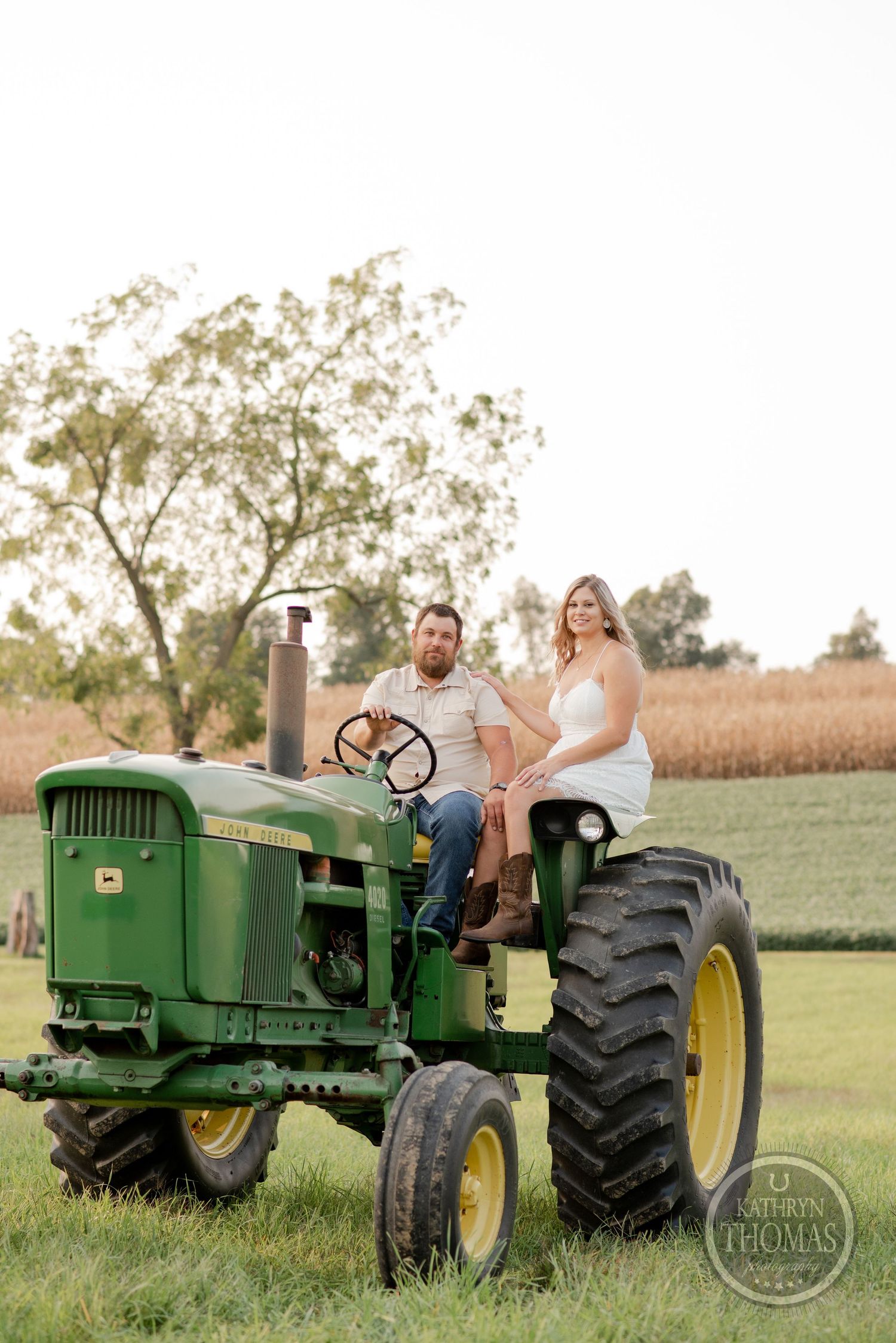 A couple sits together on a classic John Deere tractor in a rural farm setting.