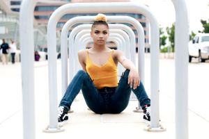 Young person in yellow tank top and jeans poses confidently in geometric white archway.