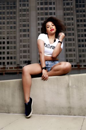 Urban fashion portrait of person in crop top and denim shorts sitting on concrete ledge.
