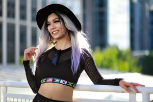 A stylish model in a black crop top and wide-brimmed hat poses against an urban backdrop with purple-tinted hair.