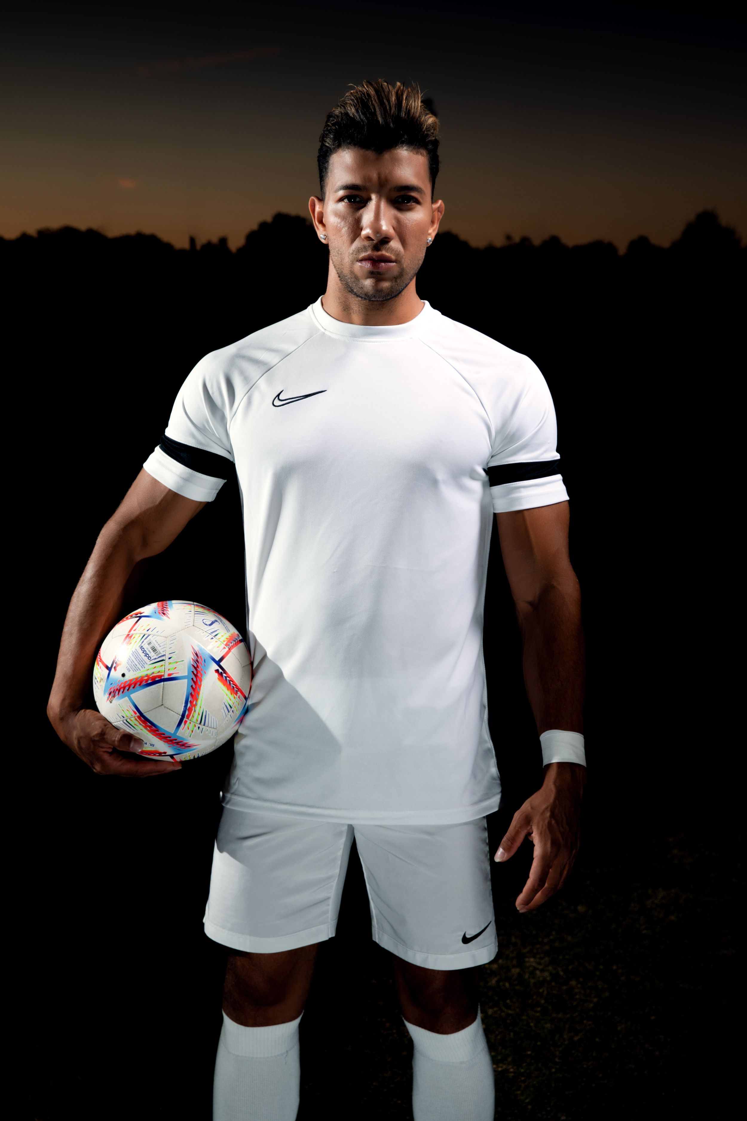 Soccer player in white Nike uniform holding ball against sunset background.