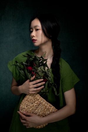 Classic portrait in emerald green dress holding flowers and woven basket against dark backdrop.