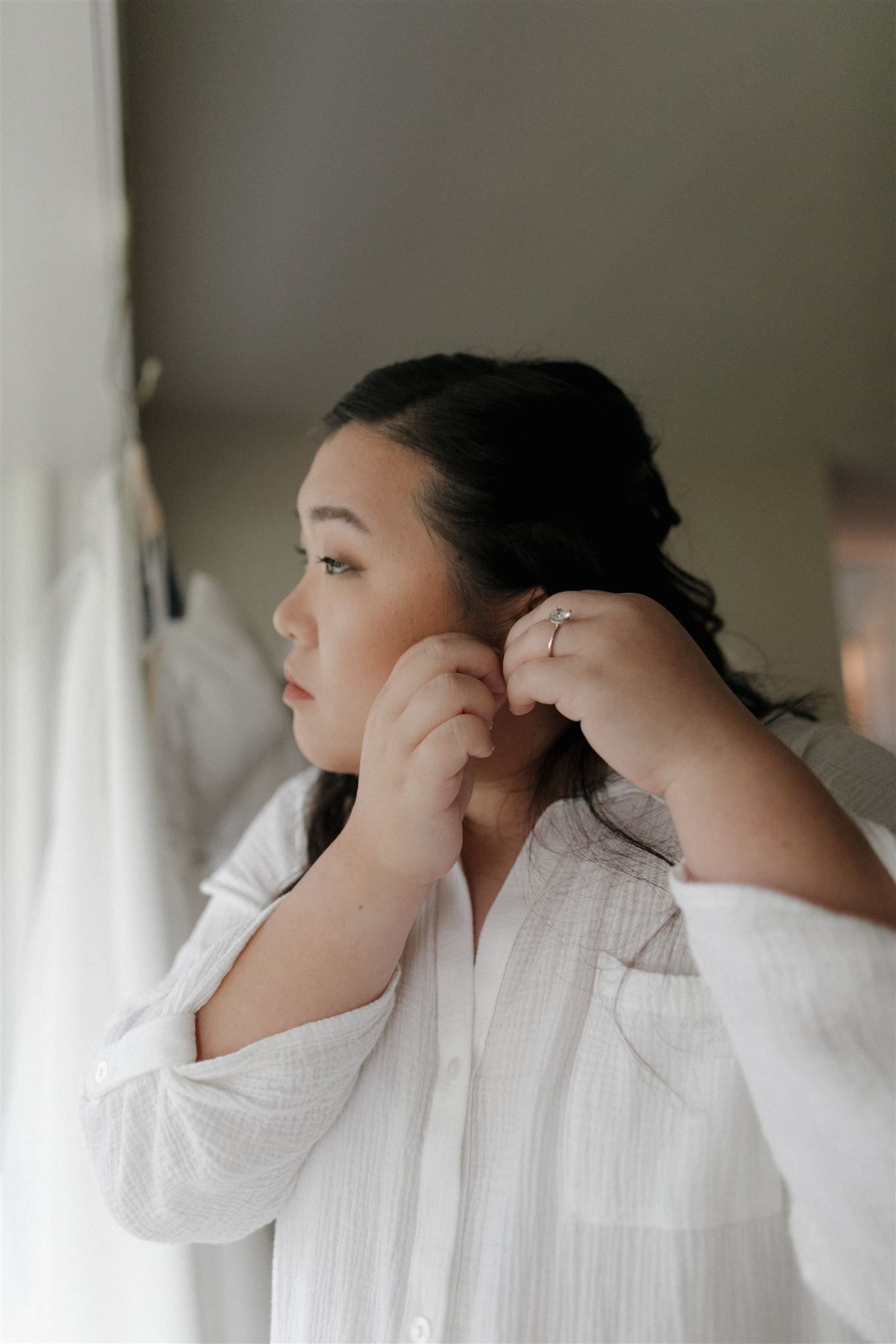 A person in white adjusts their earrings while gazing out a window.