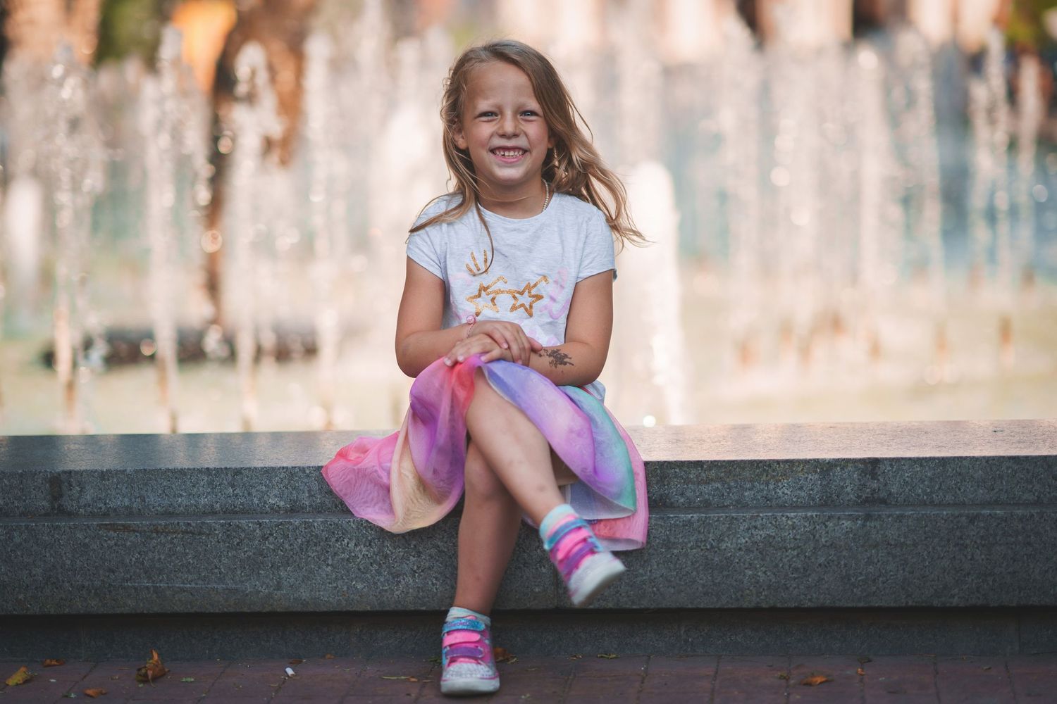 Child sitting on stone ledge by fountain wearing white t-shirt and colorful skirt.