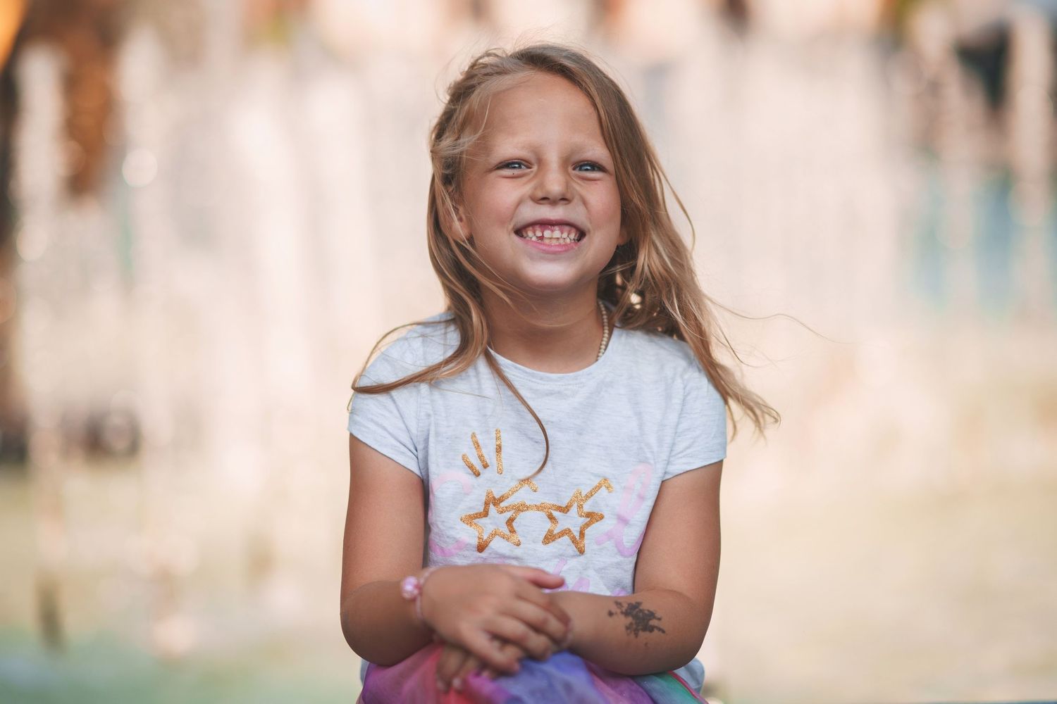 Child in white t-shirt with gold star design sitting and smiling near a fountain.