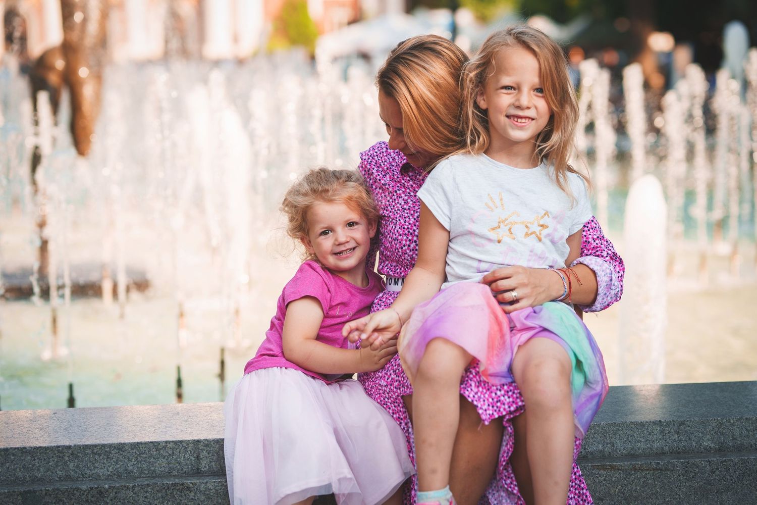 Children sitting together by fountain wearing pink and purple dresses.