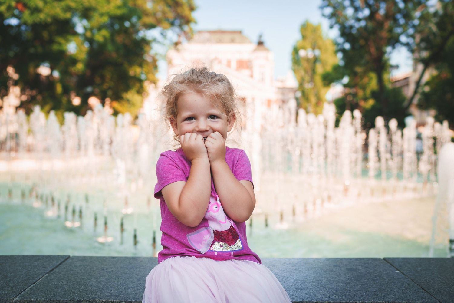 Child in pink dress covering mouth while sitting by fountain with playful expression.