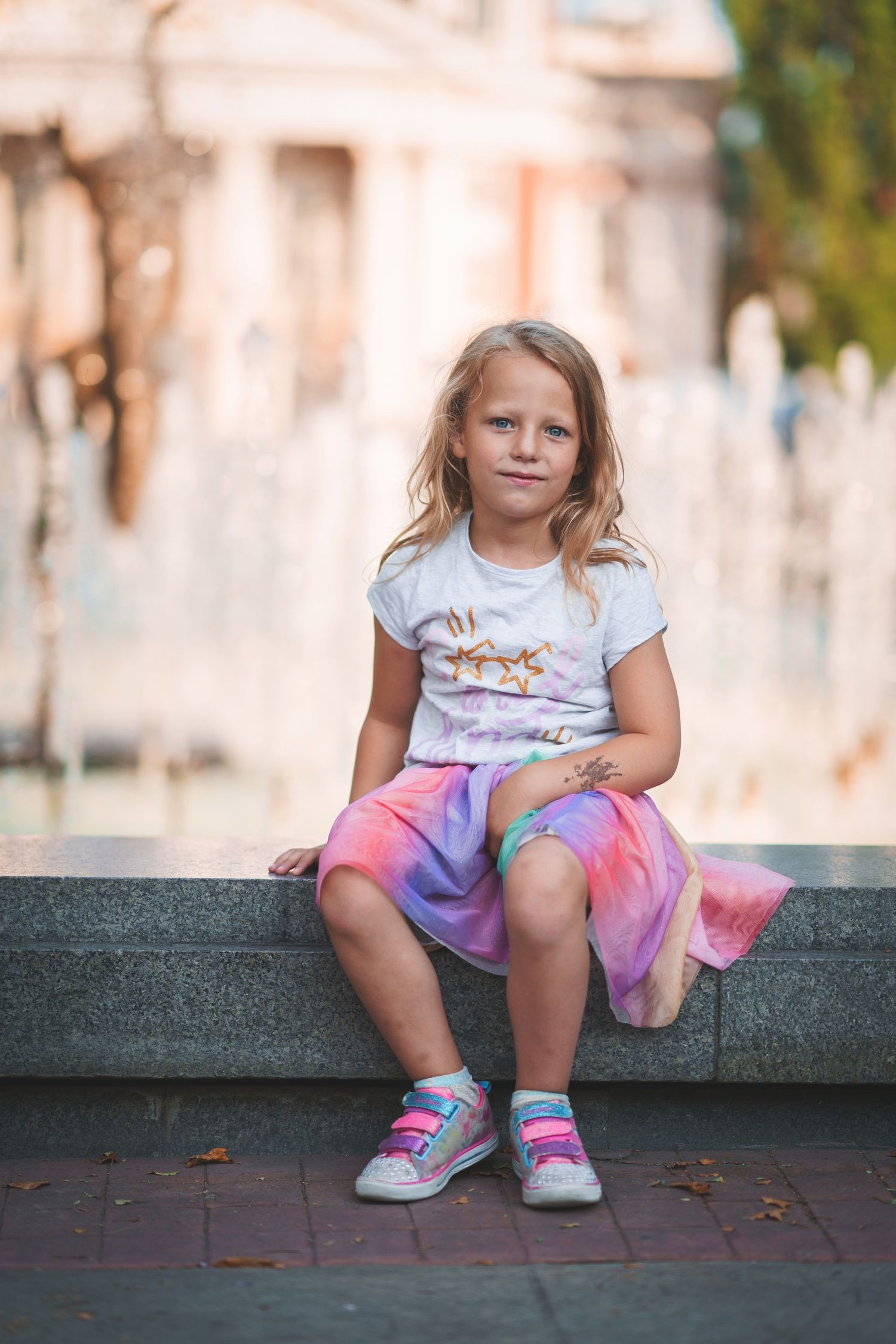 Child in casual attire sitting thoughtfully on stone ledge near fountain.