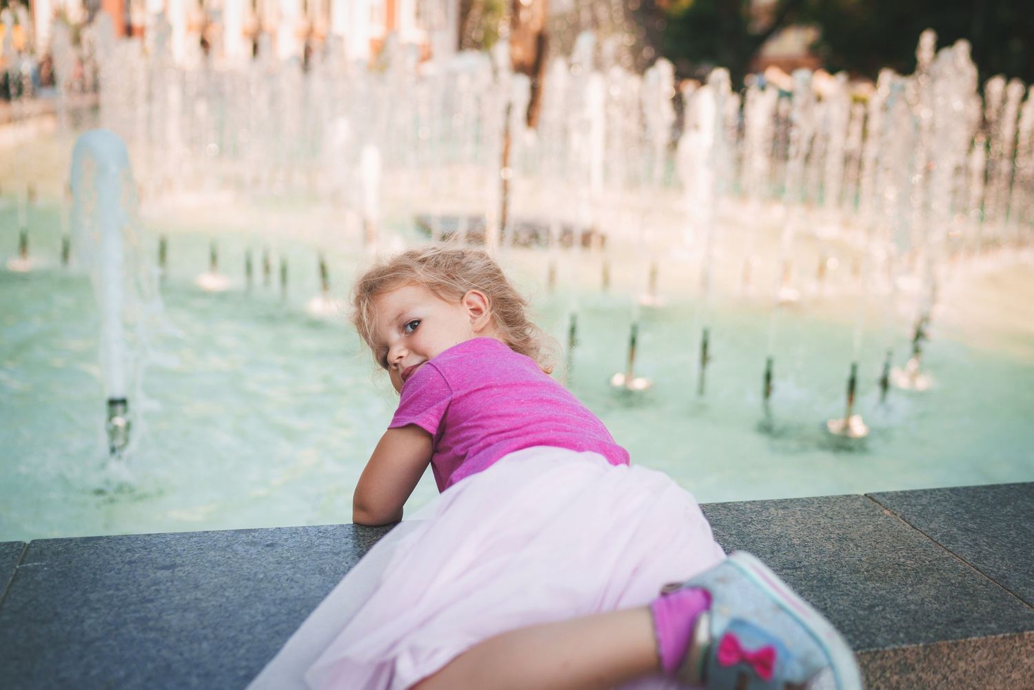 Child in pink shirt leaning over stone ledge near fountain jets.