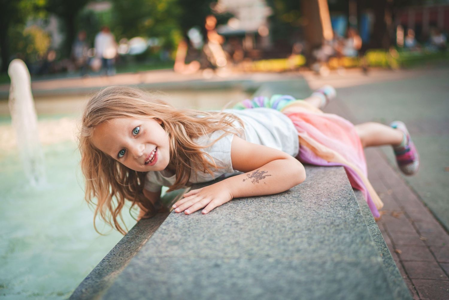 Child in light colored dress lying playfully near a fountain on a warm summer day.