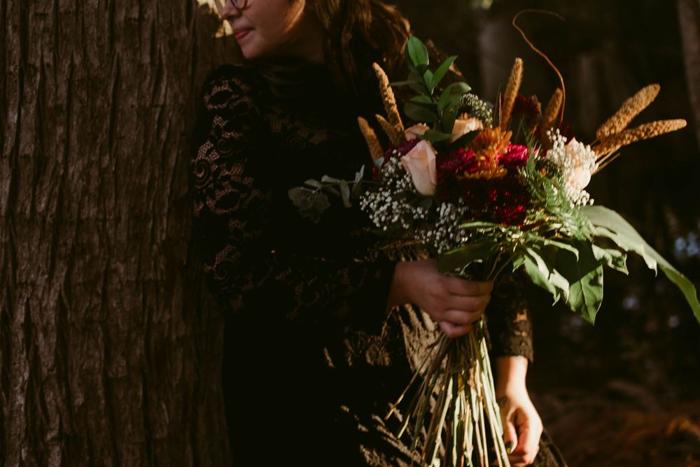 Person in black lace holding a vibrant autumn bouquet with wheat stalks against tree trunk.
